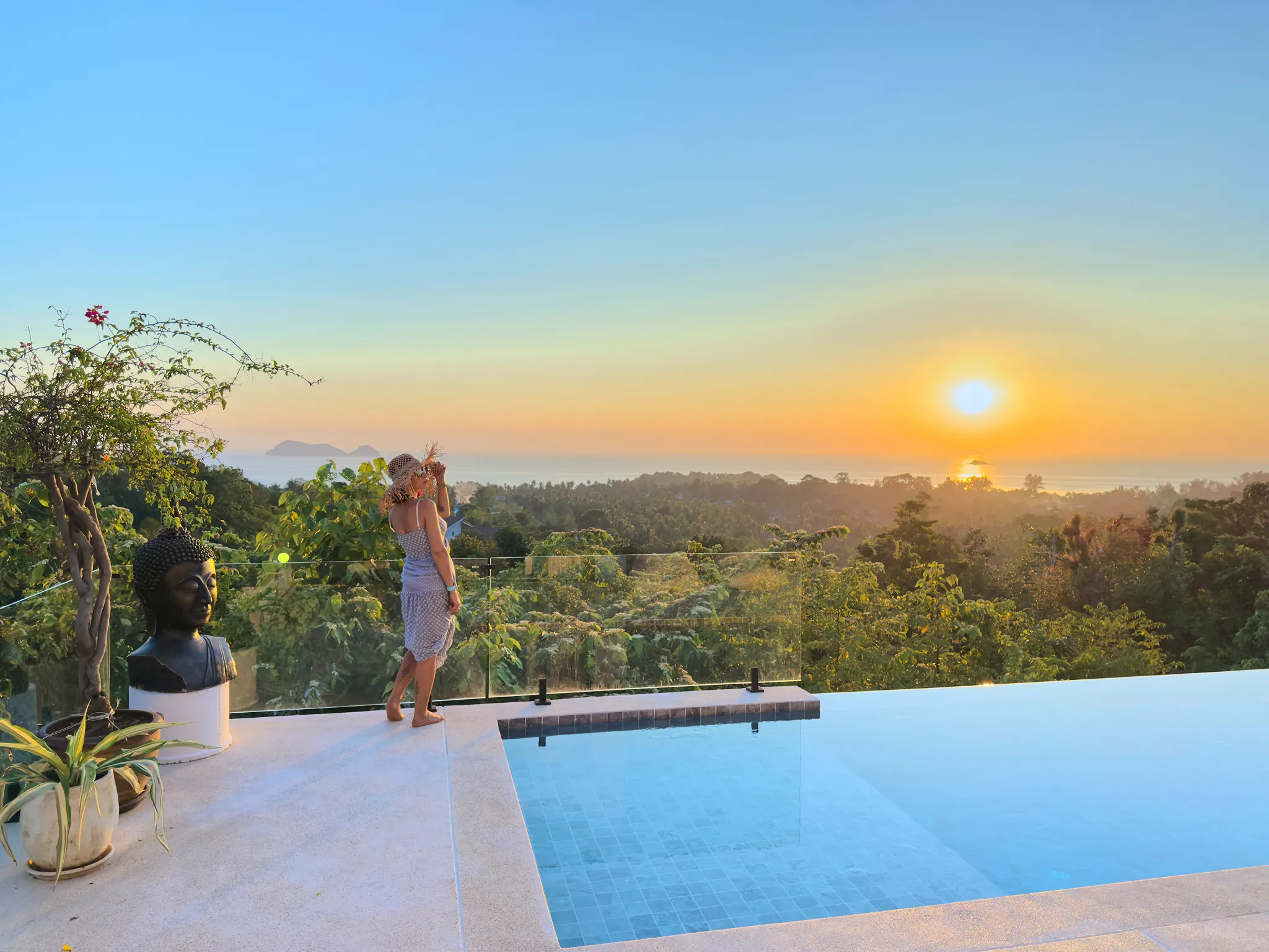 Frau in Sonnenhut und Sommerkleid steht neben einem Pool mit Glasgeländer, blickt auf die untergehende Sonne über bewaldeter Landschaft und Meer.