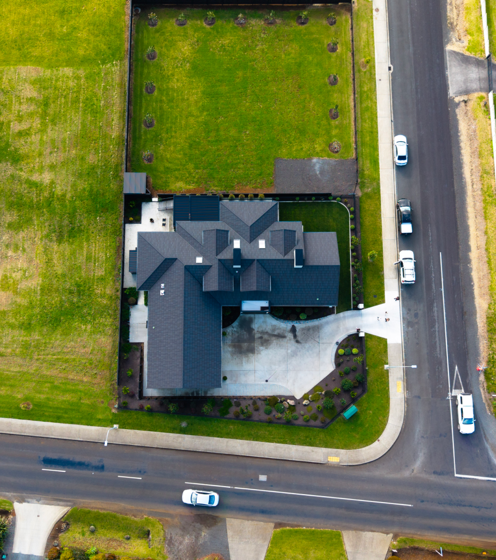 Aerial view of a large house with dark roof, driveway, and lawn at corner of two streets with cars.