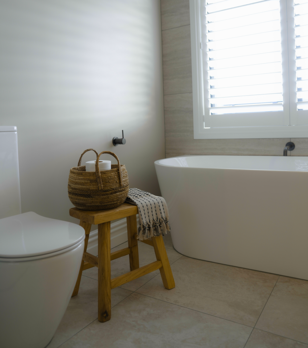 Minimalist bathroom with white bathtub, toilet, wooden stool, basket with toilet paper, and towel.