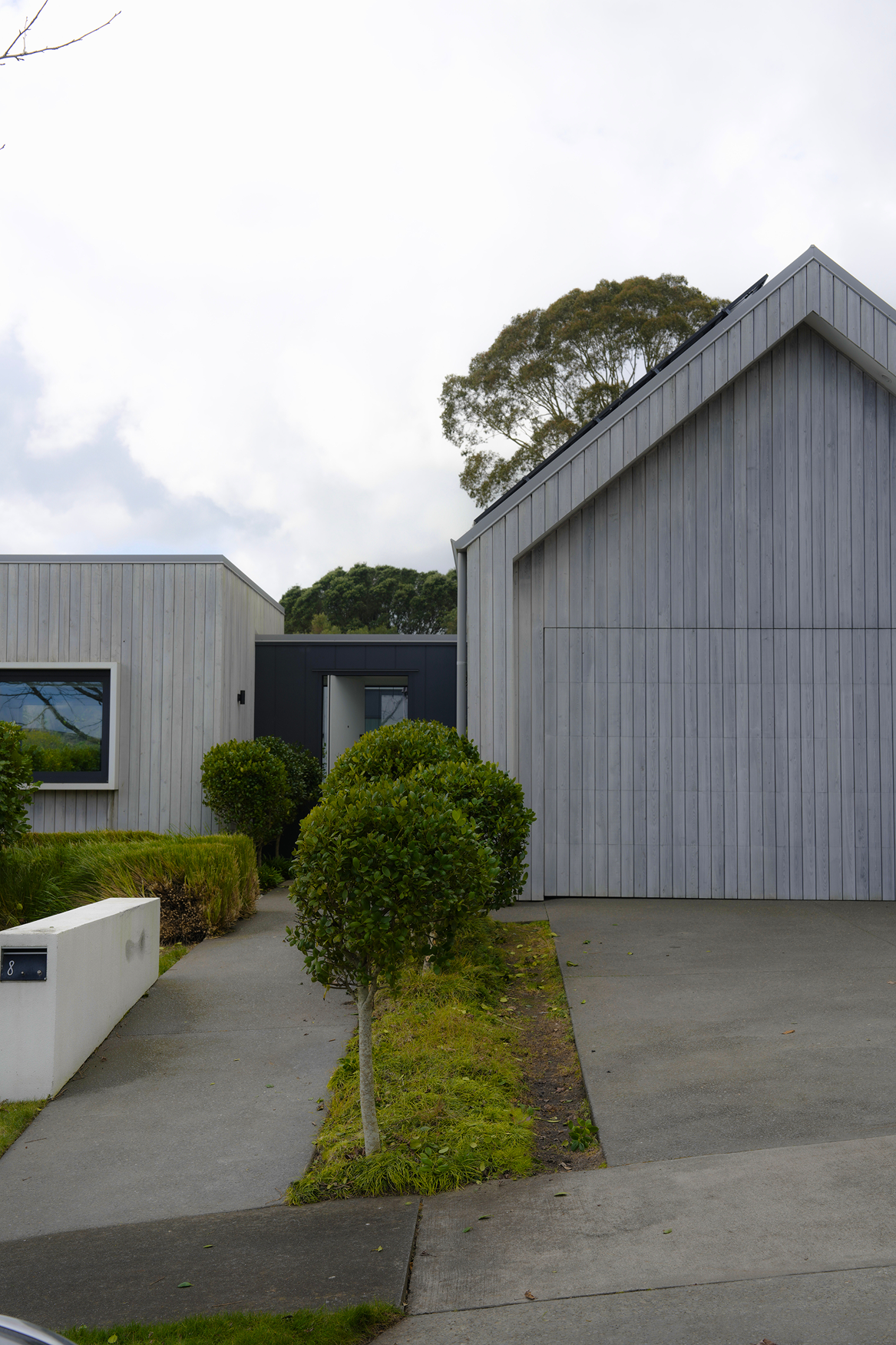 Modern house with light wood siding, trimmed bushes, concrete driveway, and cloudy sky background.
