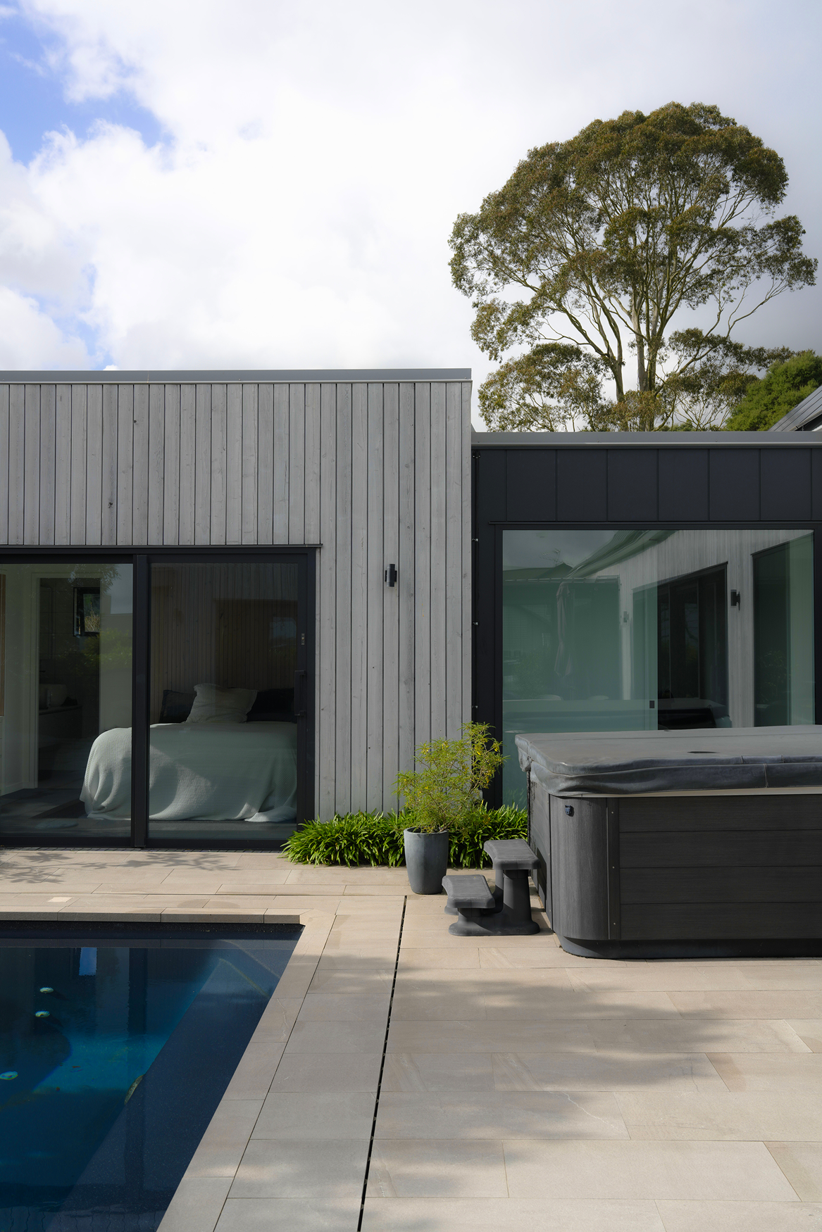 Modern backyard with a pool, hot tub, potted plant, and sliding glass doors showing a bedroom.