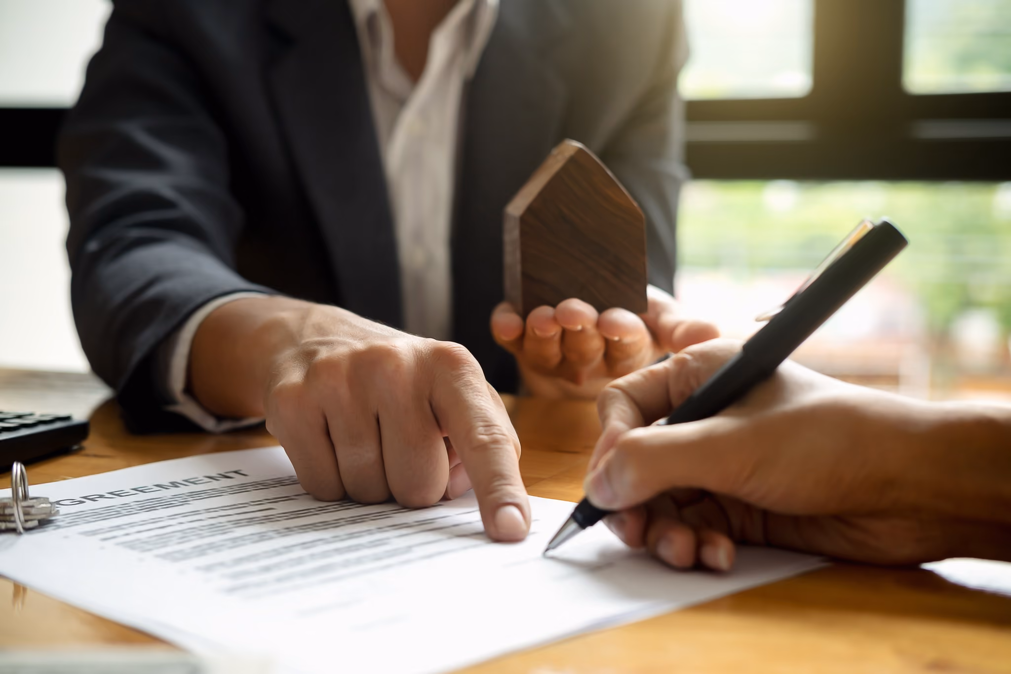 A person in a suit signing a property document while holding a small wooden house model