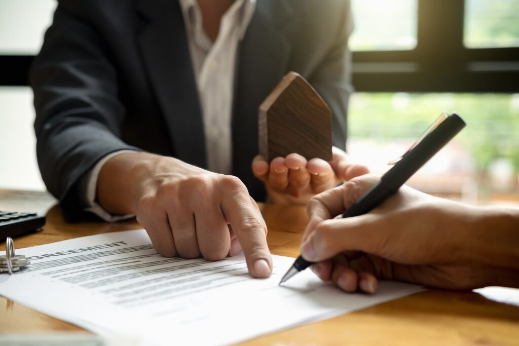 A person in a suit signing a property document while holding a small wooden house model