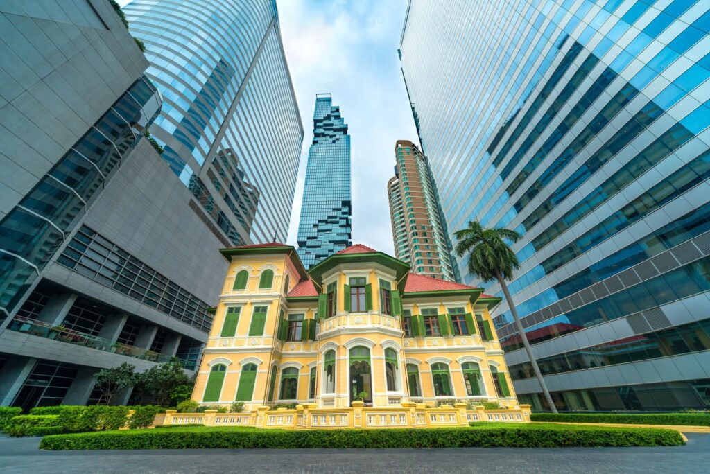 A restored yellow colonial-era building framed by modern glass office towers in central Bangkok