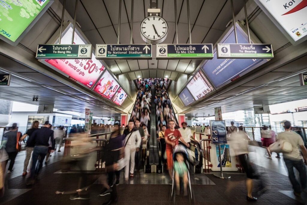 Commuters crossing a BTS station concourse in Bangkok beneath directional signage and a station clock