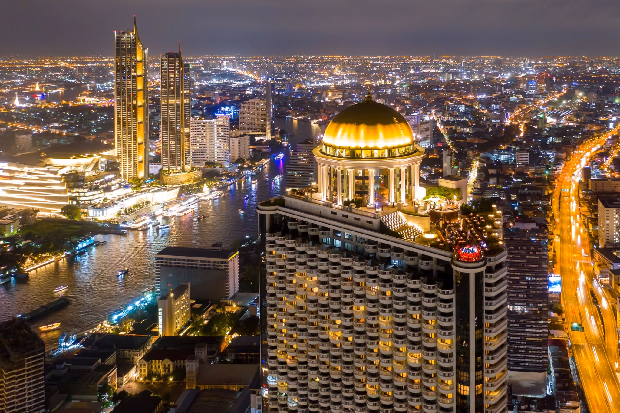 The Bangkok riverside at night, with lit towers and the gold dome of State Tower above the Chao Phraya