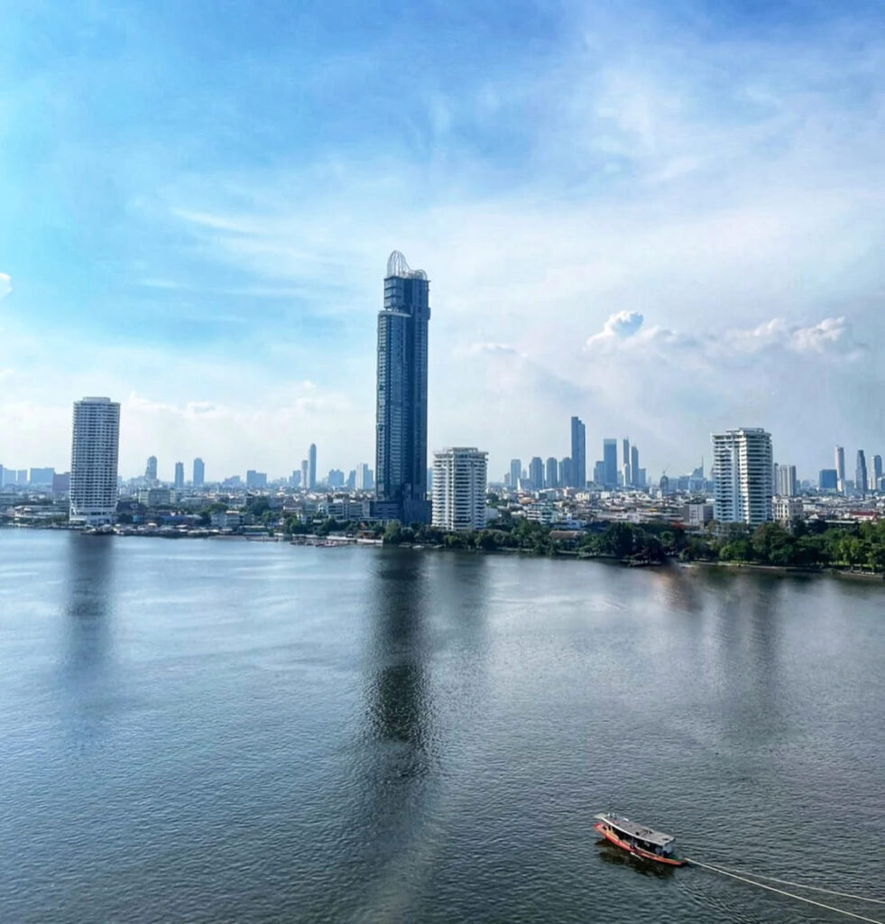 The Canapaya Residences tower on the Chao Phraya, with a boat passing on the water below