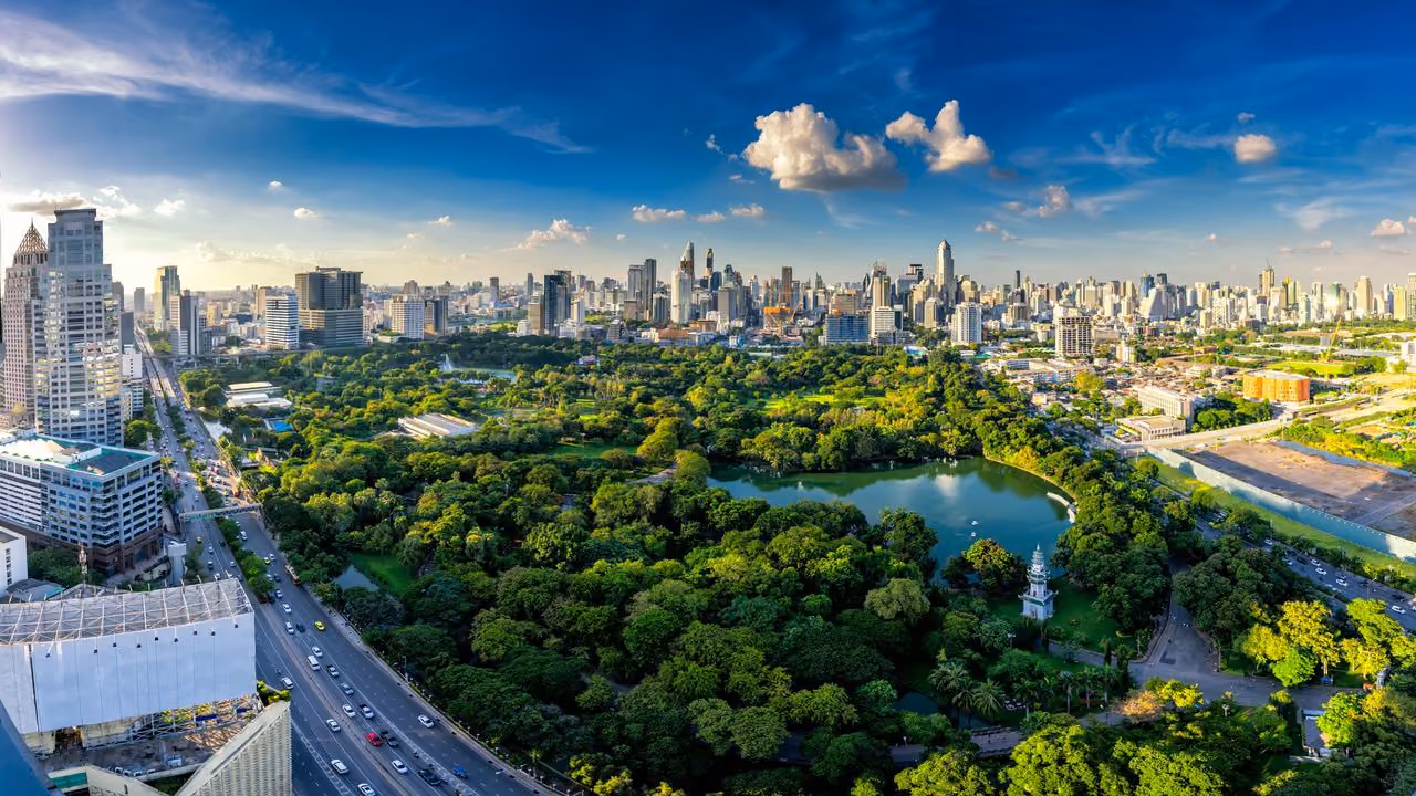 An aerial view over Lumpini Park, its lake surrounded by trees and the Bangkok skyline beyond