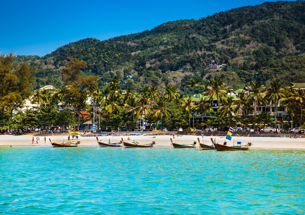 A Phuket beach with longtail boats moored on turquoise water below a forested headland