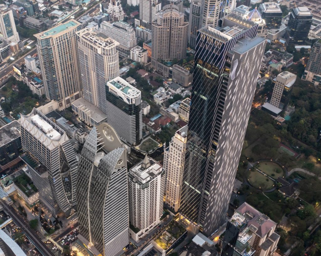 Aerial view of a tall dark tower among dense Bangkok office blocks at dusk