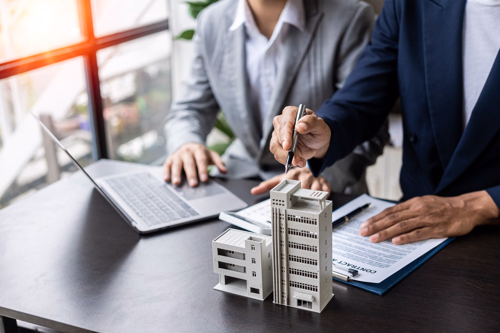 Two people in suits reviewing a contract beside small white models of apartment buildings