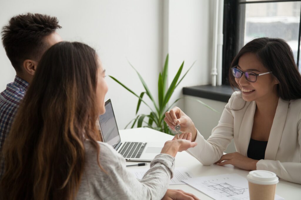 Smiling agent in glasses handing a set of keys across a desk to a young couple