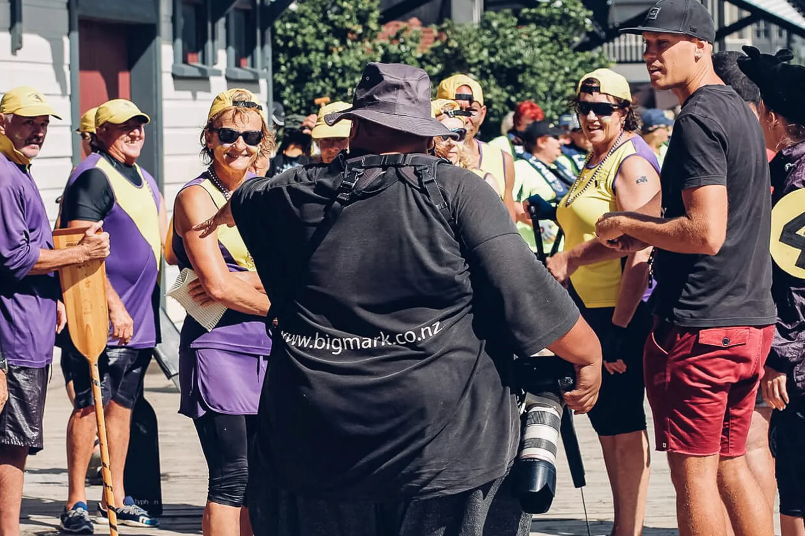 Photographer carrying a telephoto camera while speaking with a group in yellow and purple sports clothing.