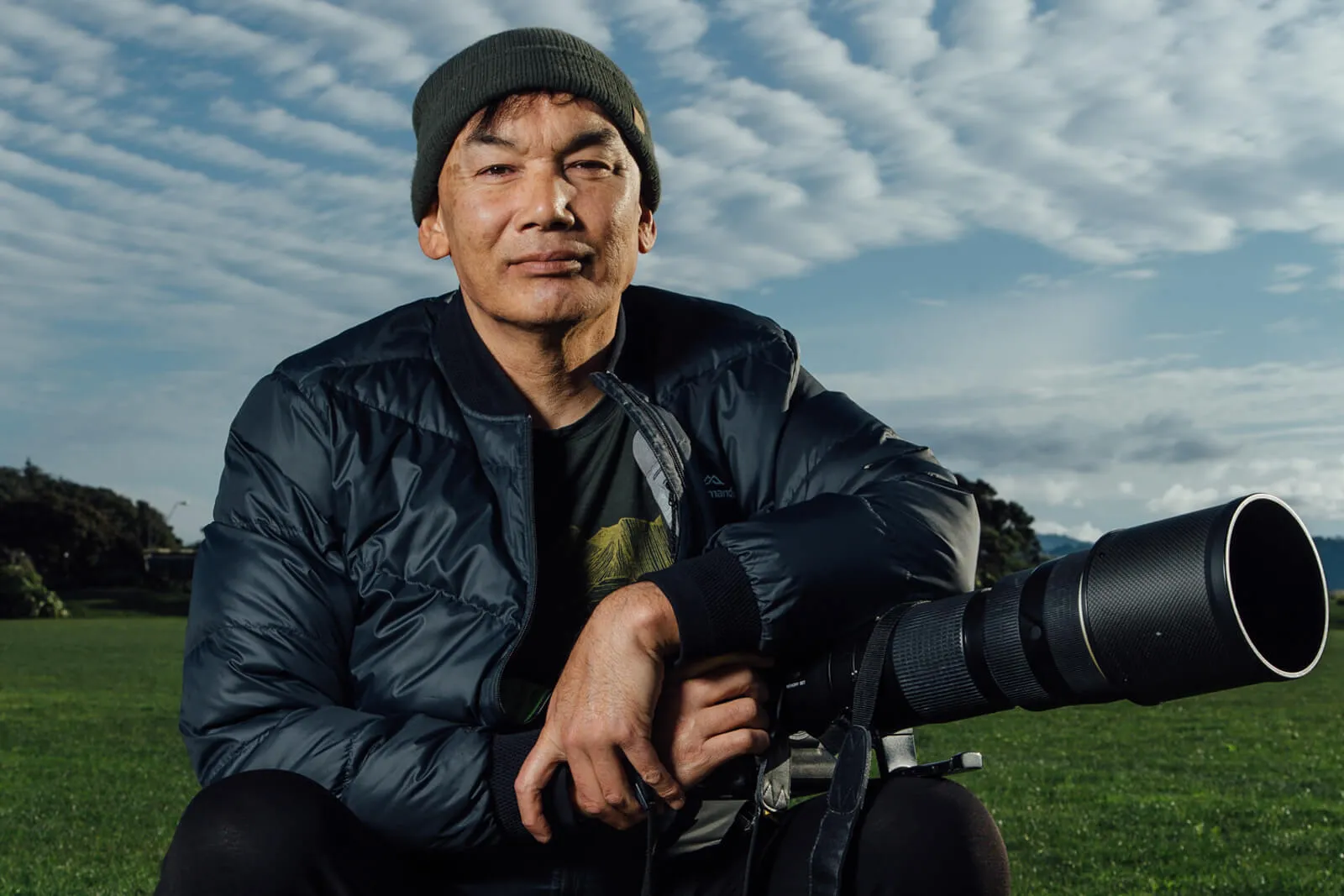 Photographer in a black beanie seated beside a camera with a large telephoto lens outdoors.