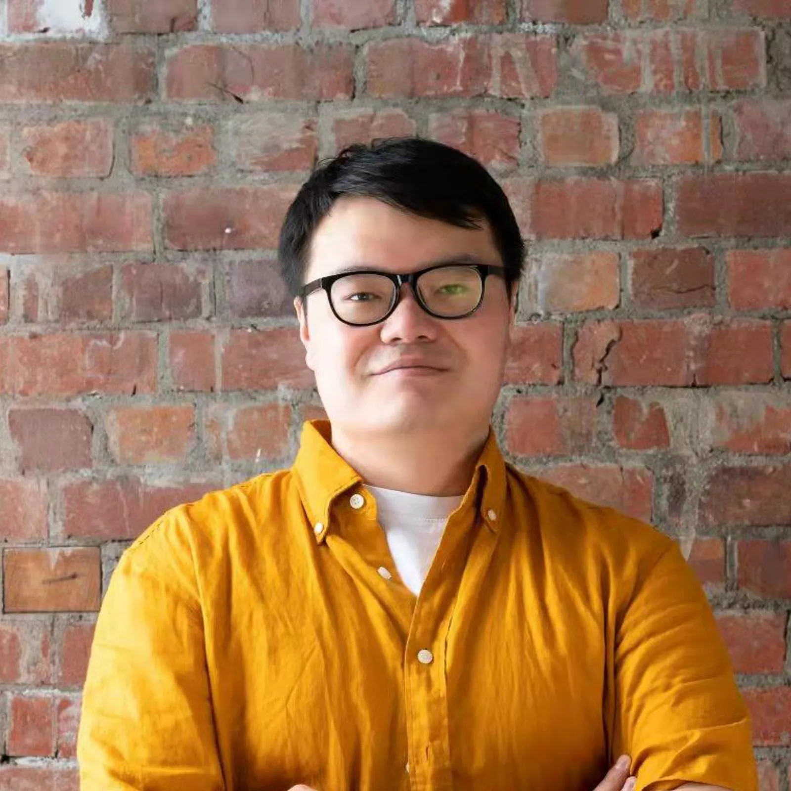 Portrait of a photographer in glasses and a mustard shirt standing against a brick wall.