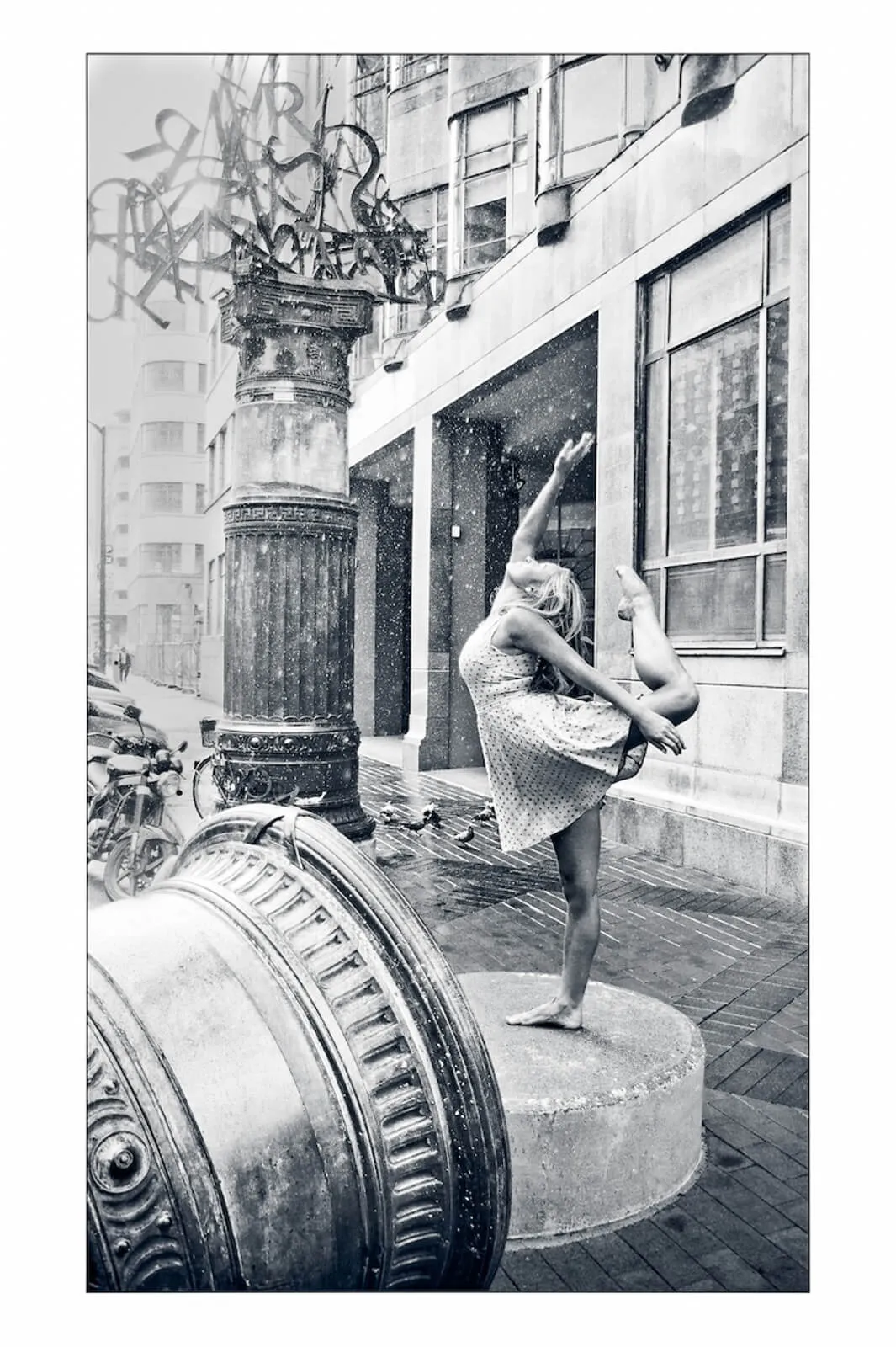 Black-and-white photograph of a dancer balancing on one leg on a city street.