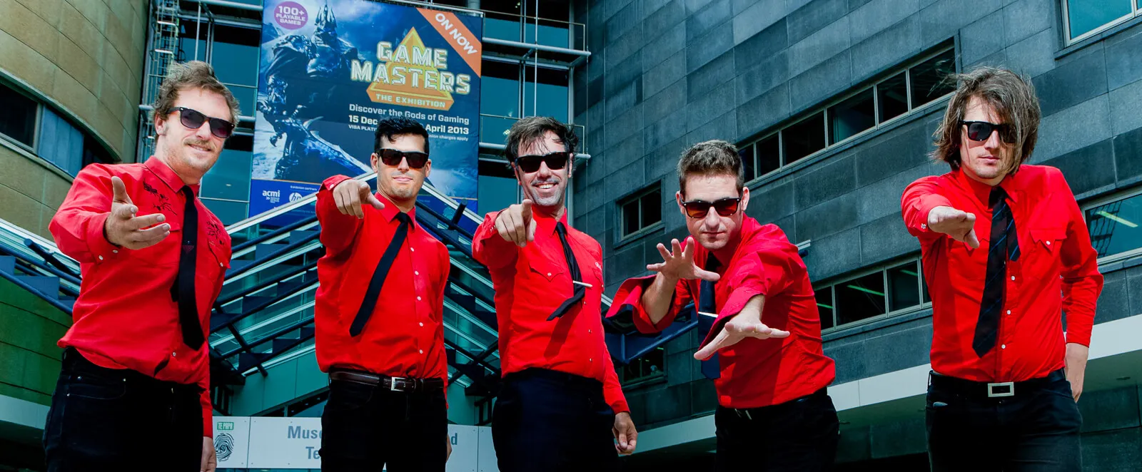 Five band members in matching red shirts and black ties reaching toward the camera.