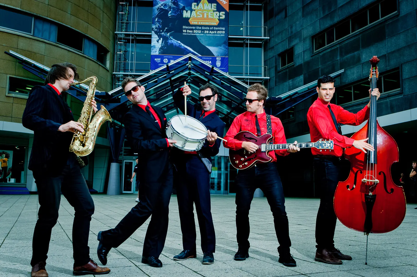 Five-piece band performing outdoors with saxophone, drums, guitar and double bass.