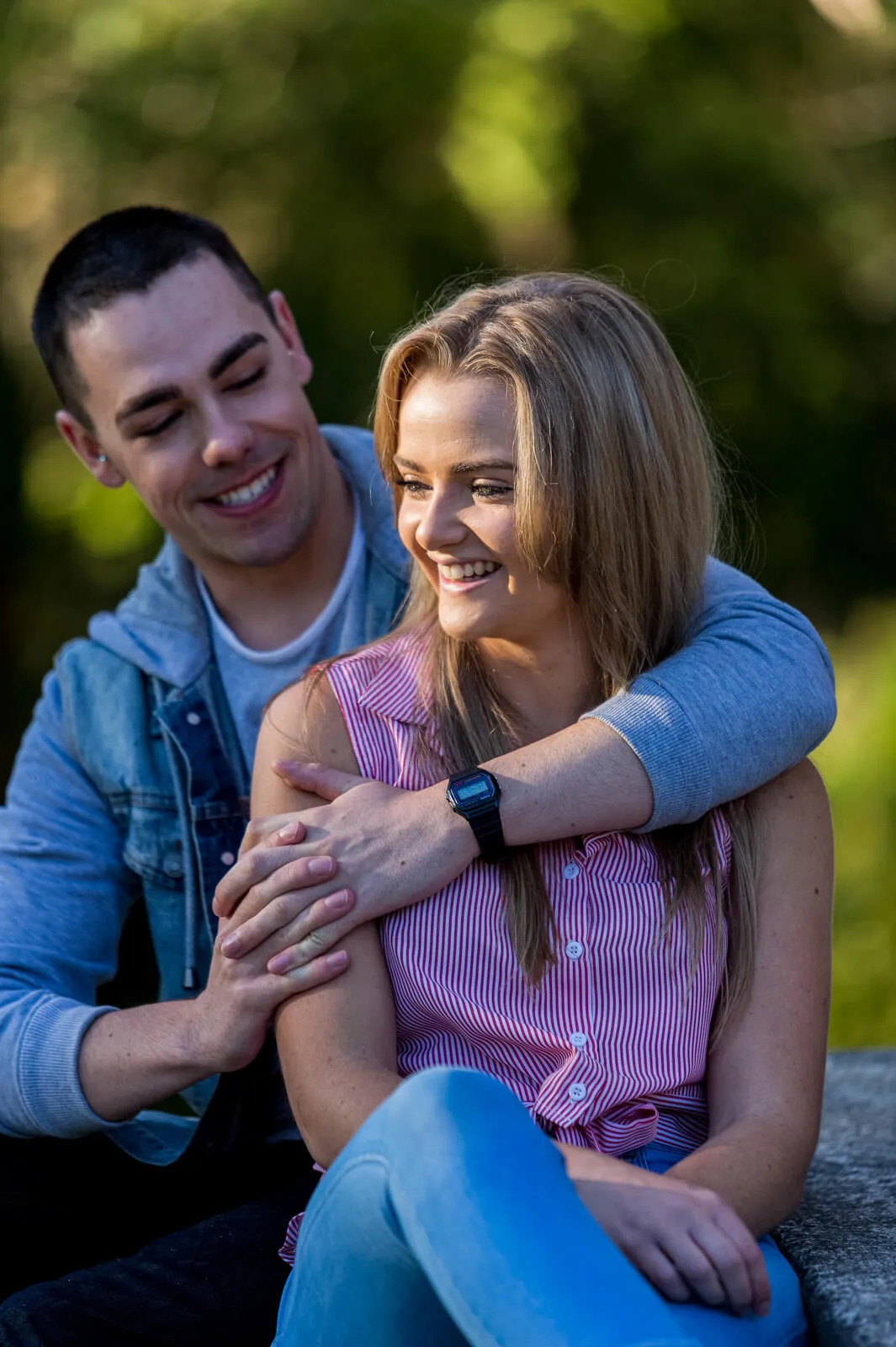 Smiling couple seated outdoors with the man embracing the woman from behind.