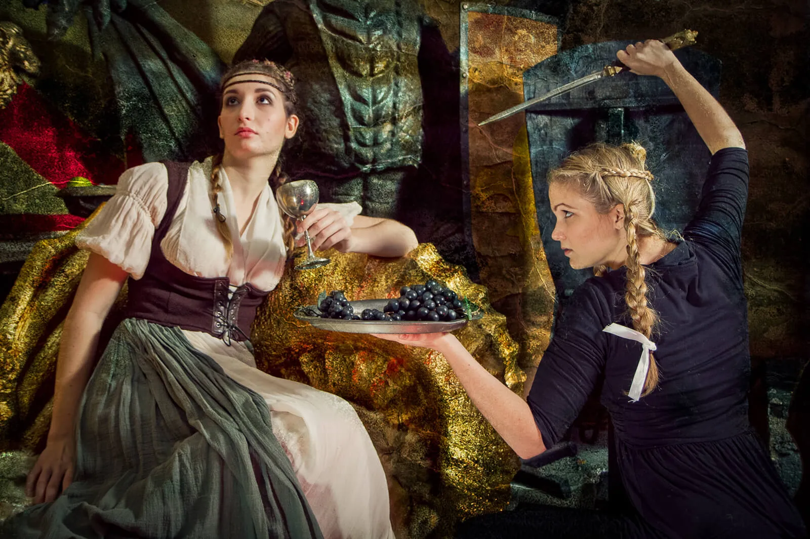 Two women in fantasy costumes on an ornate set, one presenting a platter of grapes while holding a knife overhead.