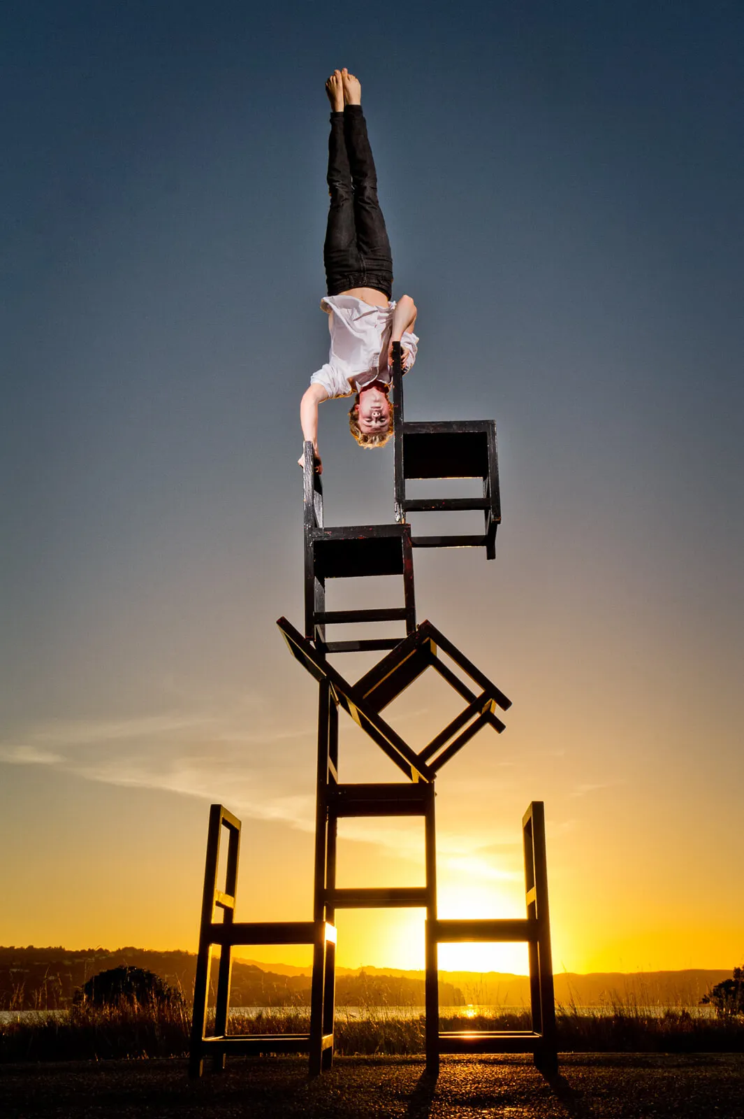 Acrobat balancing upside down on a tall stack of chairs at sunset.