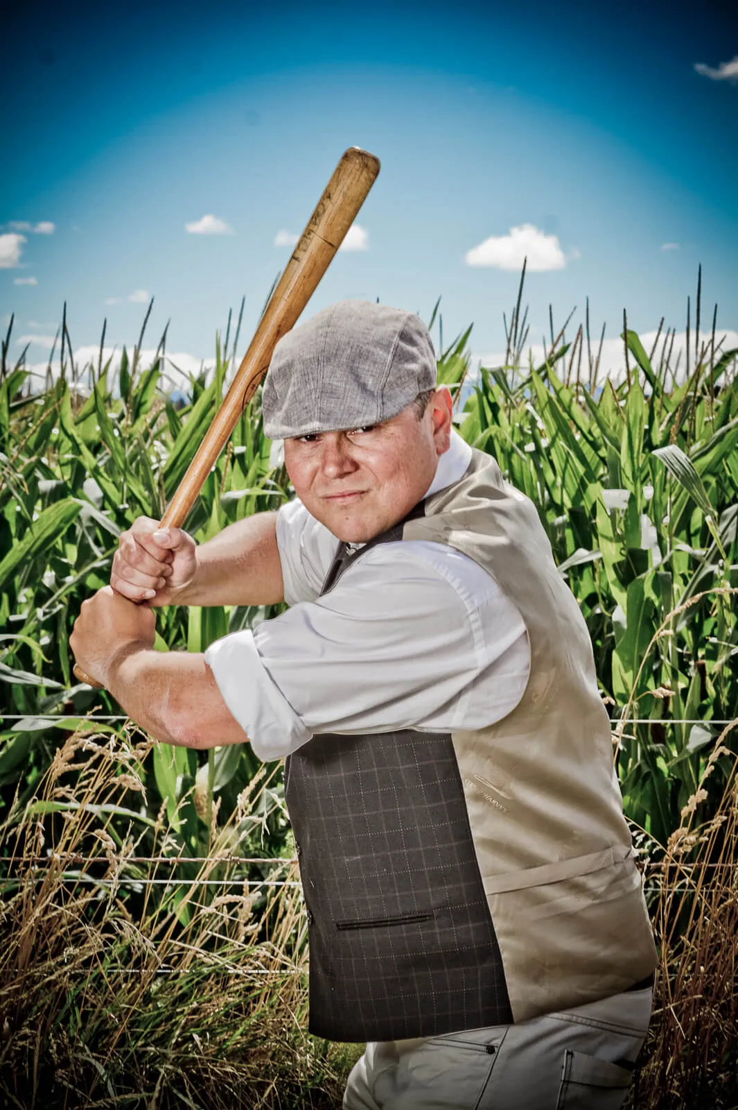 Man in a flat cap holding a cricket bat in front of a cornfield.