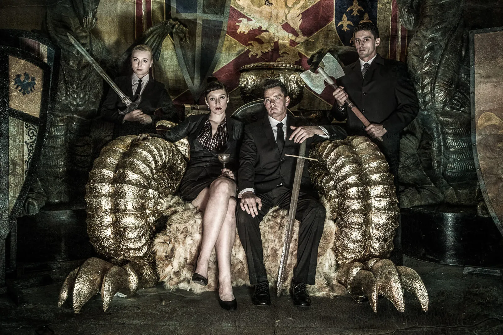 Four people in dark formal clothing posing on an ornate fantasy throne with prop weapons.