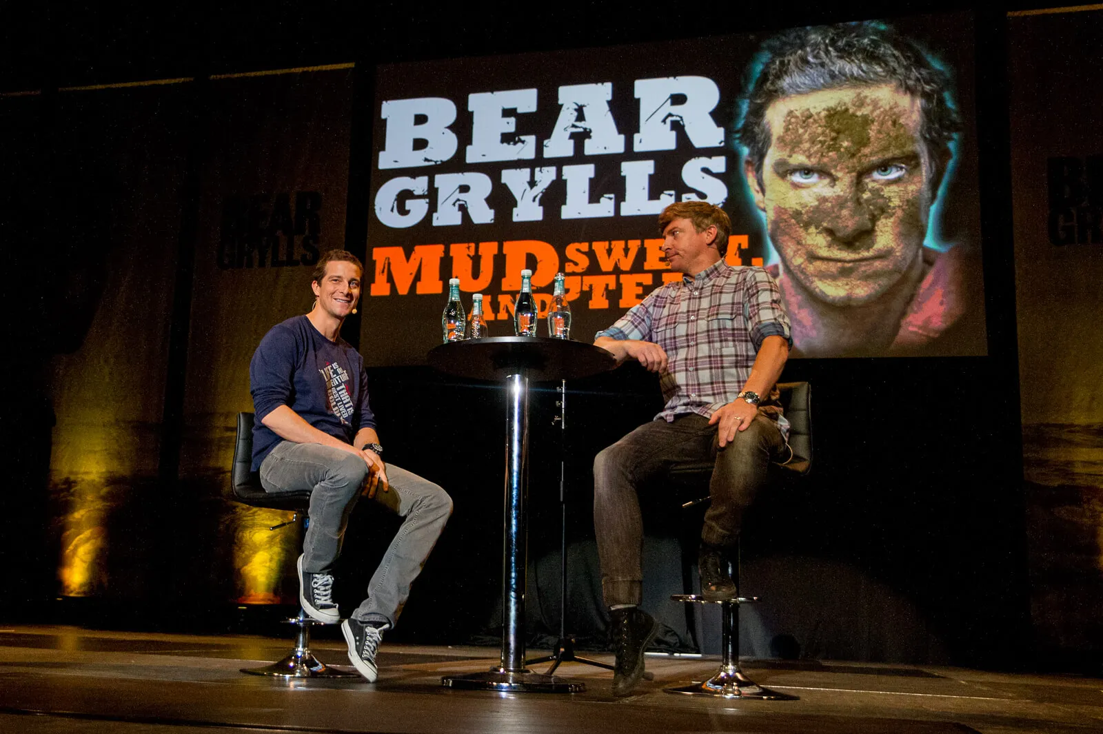 Two men seated for an interview on stage in front of a Bear Grylls event backdrop.