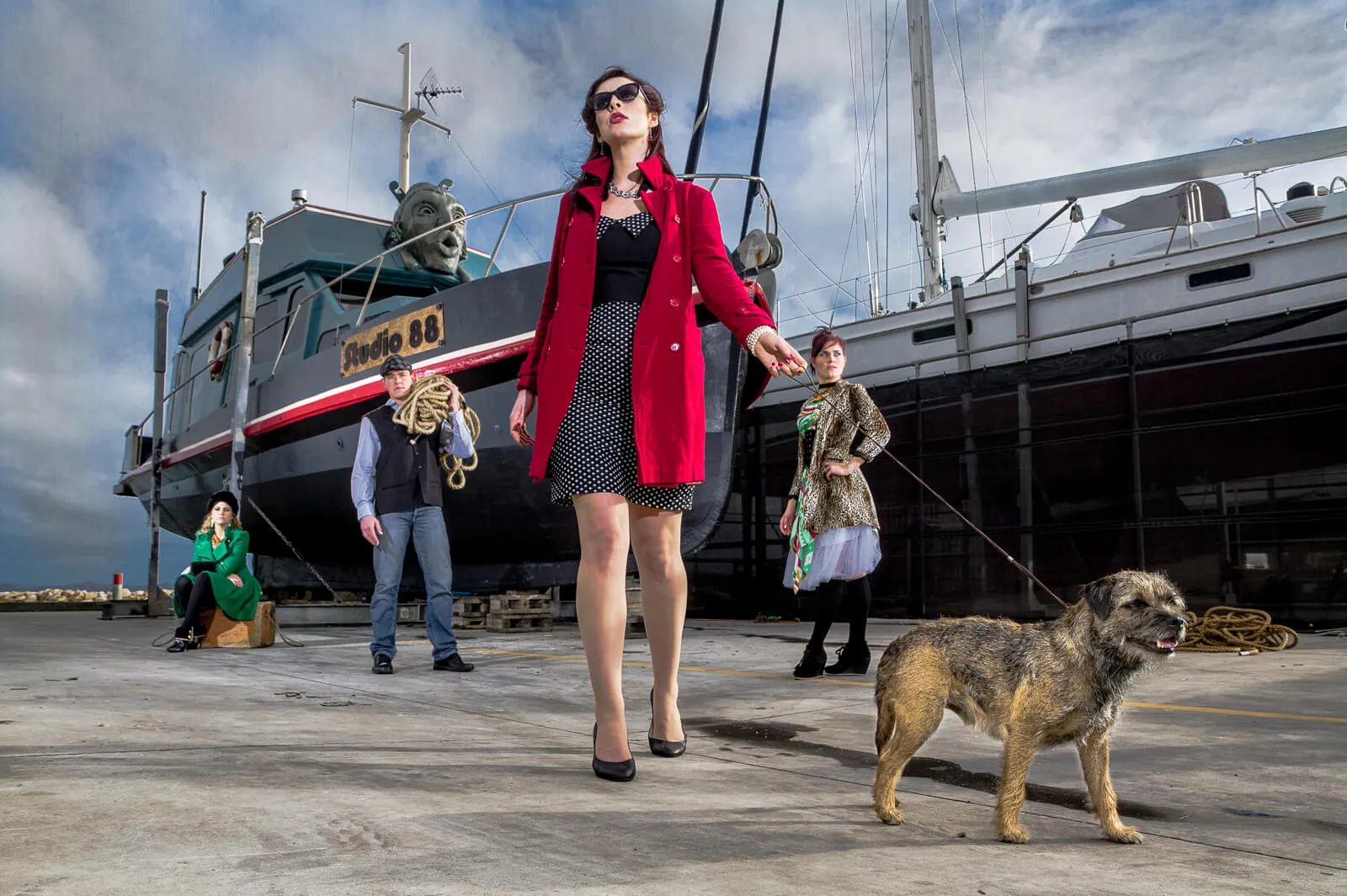 Woman in a red coat walking a small dog past people and boats in a shipyard.