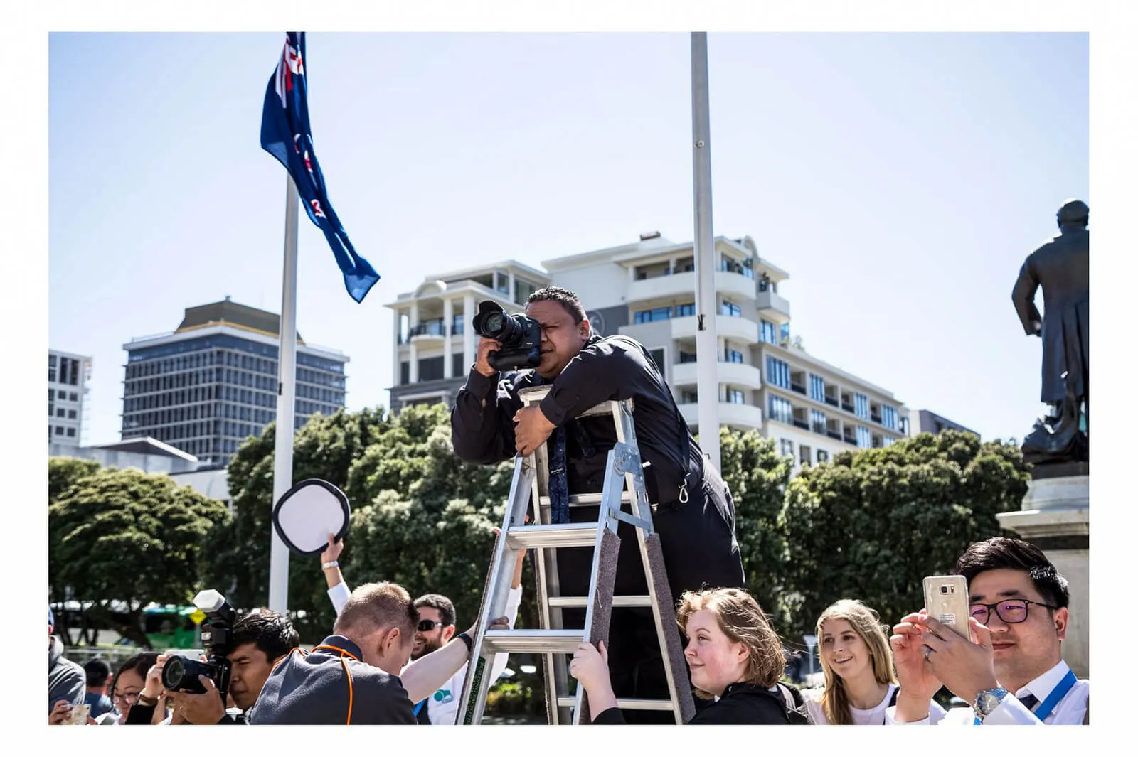 Photographer standing on a ladder taking a picture at an outdoor public event beneath a New Zealand flag.