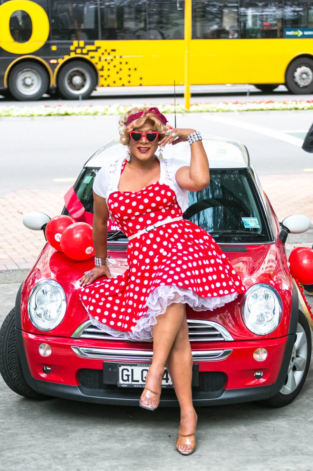 Woman in a red polka-dot dress and heart-shaped sunglasses sitting on a red car decorated with balloons.