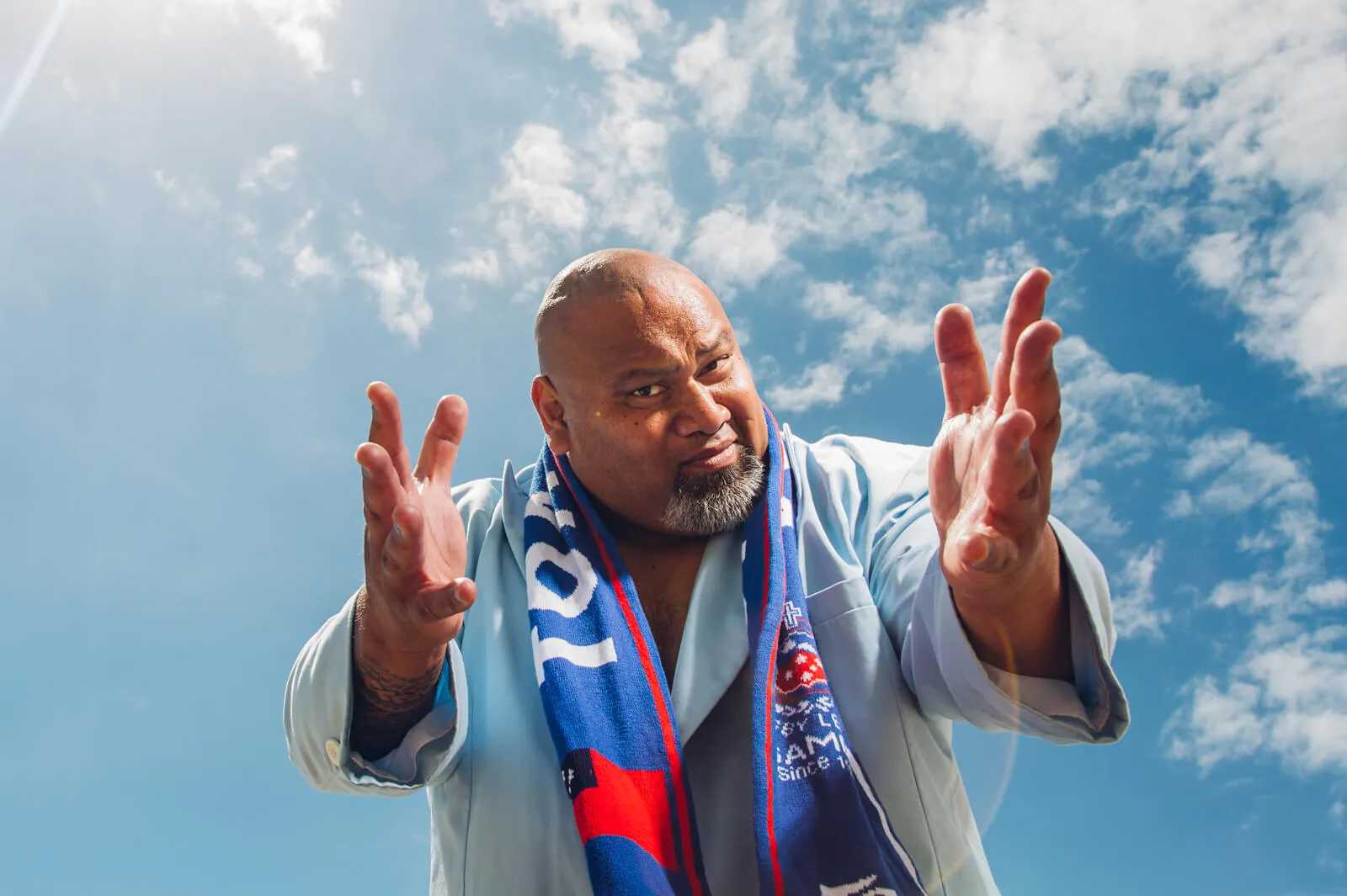 Man wearing a blue and red sports scarf reaching both hands toward the camera beneath a bright sky.
