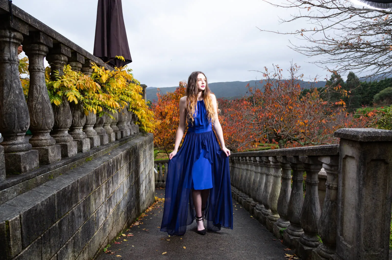 Woman in a long blue dress standing between stone balustrades in an autumn garden.