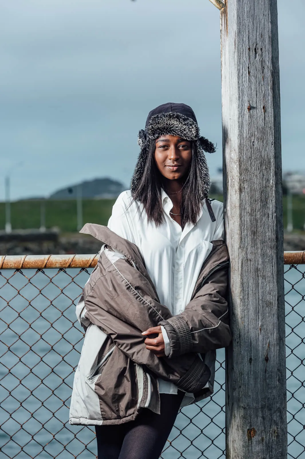 Person in a winter hat and oversized coat standing beside a waterfront fence.