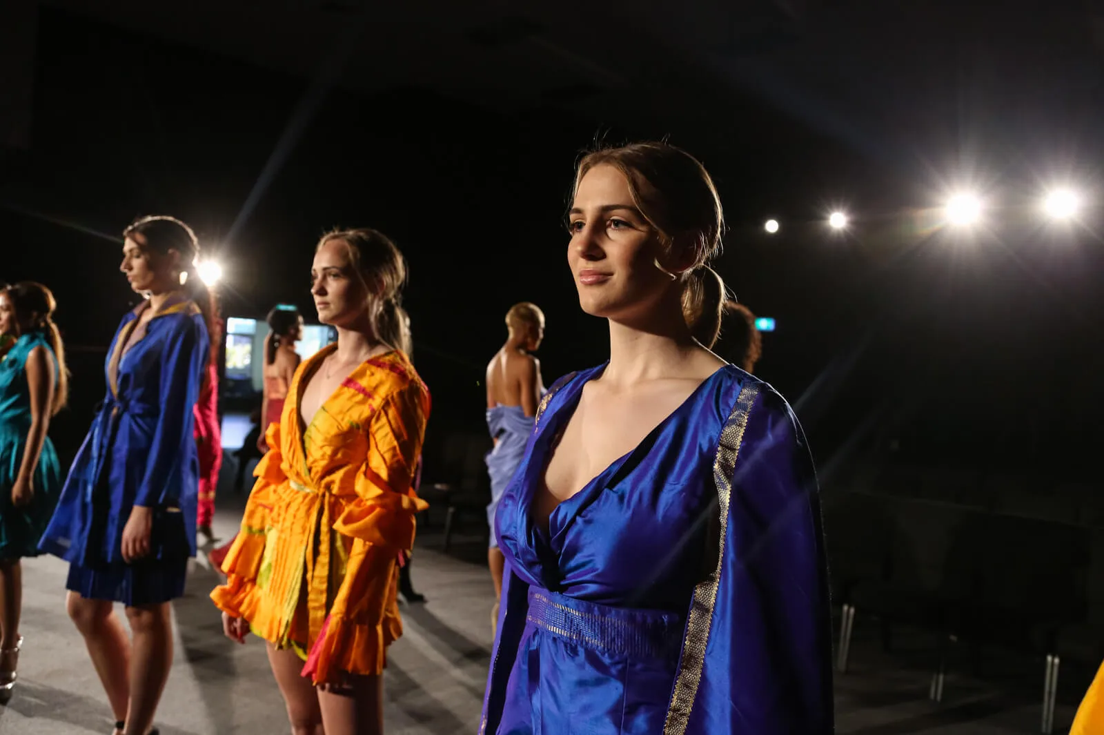 Models in bright dresses waiting backstage under stage lights.