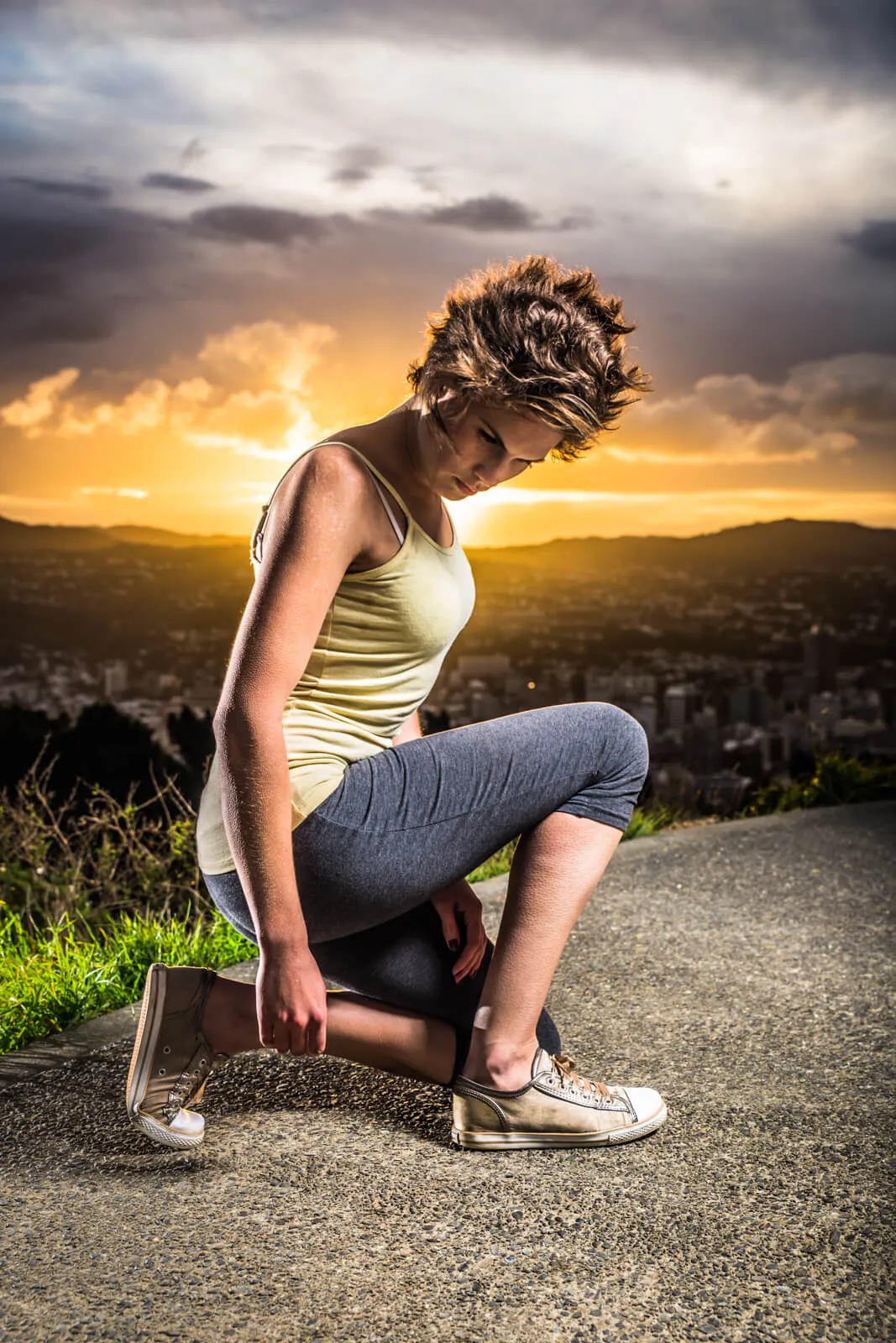 Person kneeling on a hillside path at sunset with a city in the background.