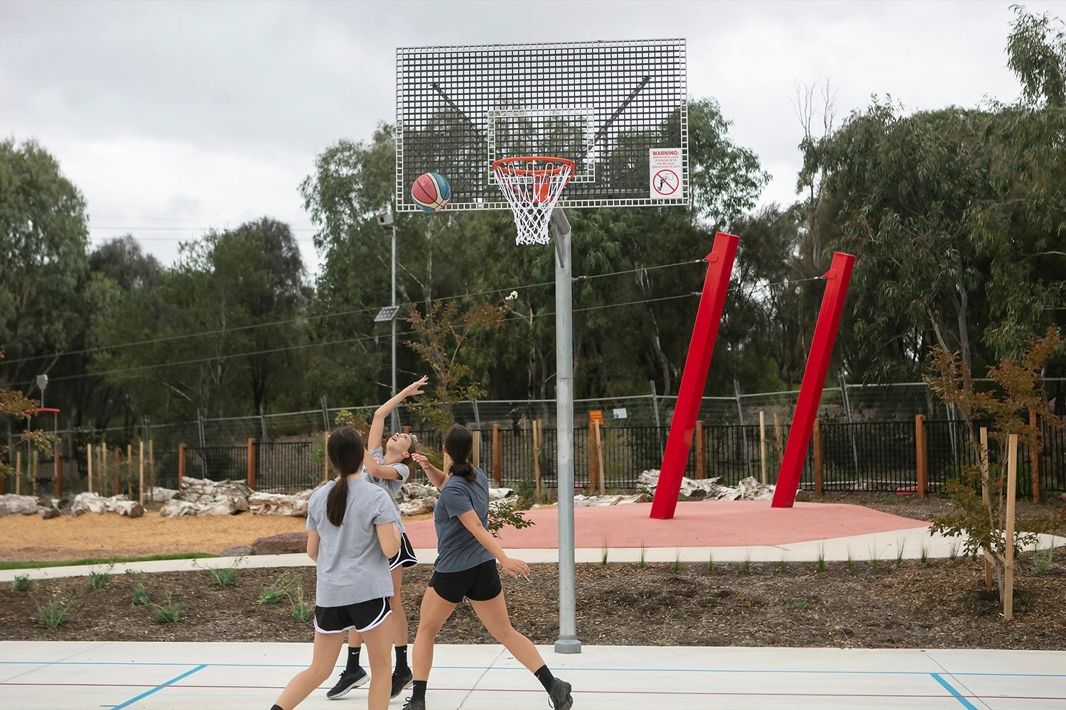 Three girls playing basketball outdoors, one shooting the ball towards the hoop on a cloudy day.