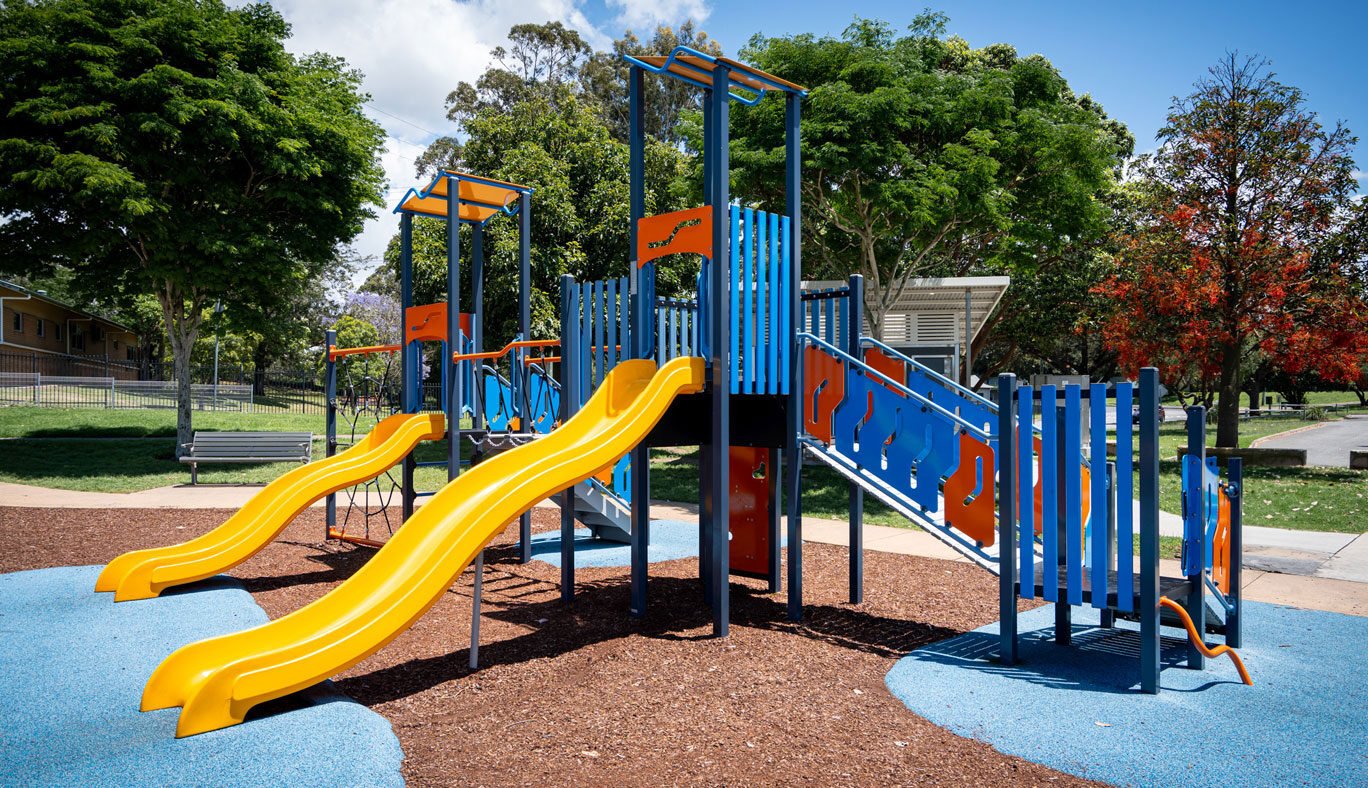 Children's playground with two yellow slides, blue and orange climbing structures, and surrounding trees.