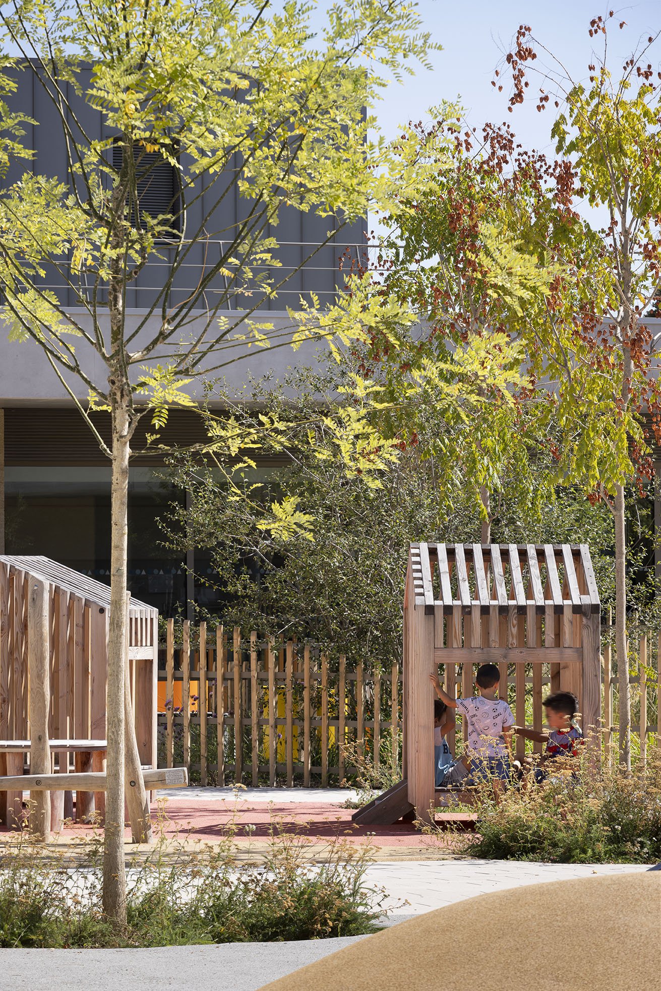 Children playing inside wooden playhouse structures at a sunny outdoor playground with trees and plants.