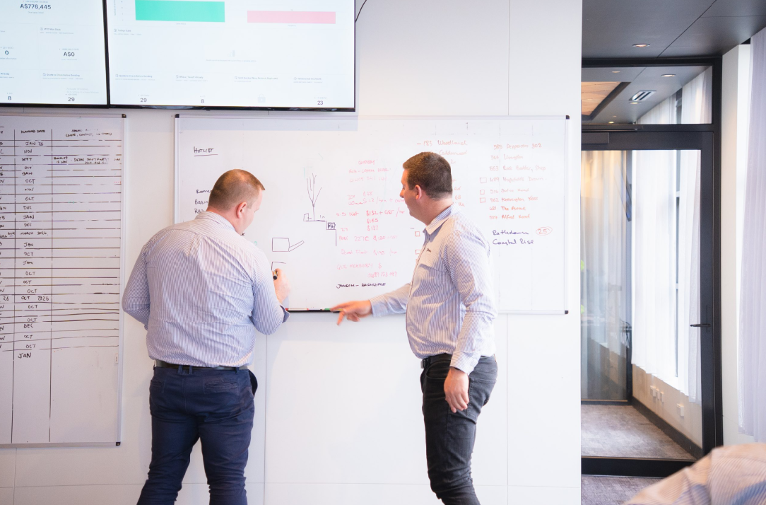 Two men in striped shirts collaborating and writing on a whiteboard with notes and diagrams.