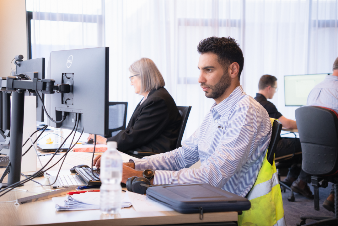 Man in striped shirt working at a desk with dual monitors and office coworkers in background.