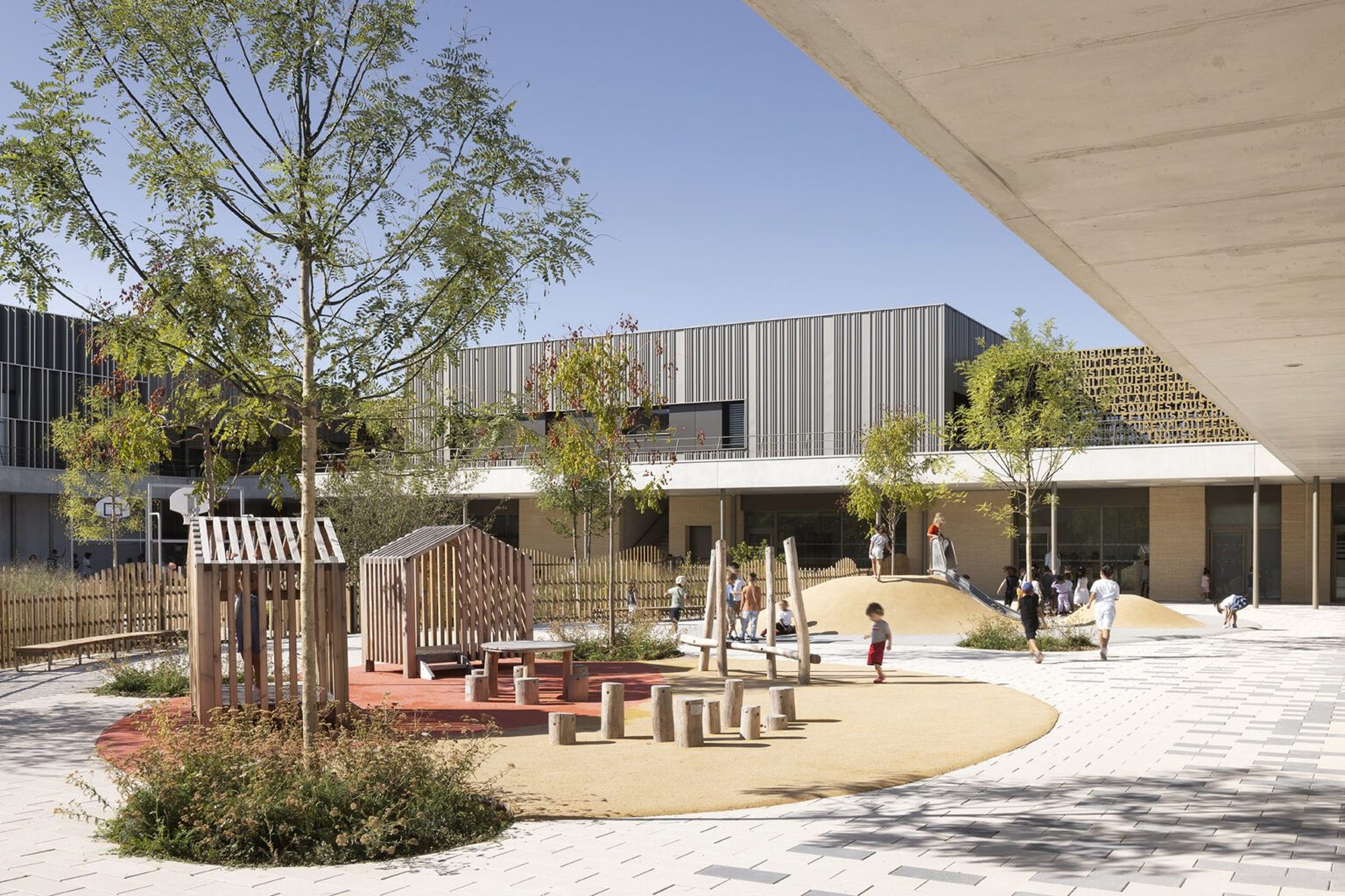Modern school playground with wooden play structures, children playing, and a two-story building in background.