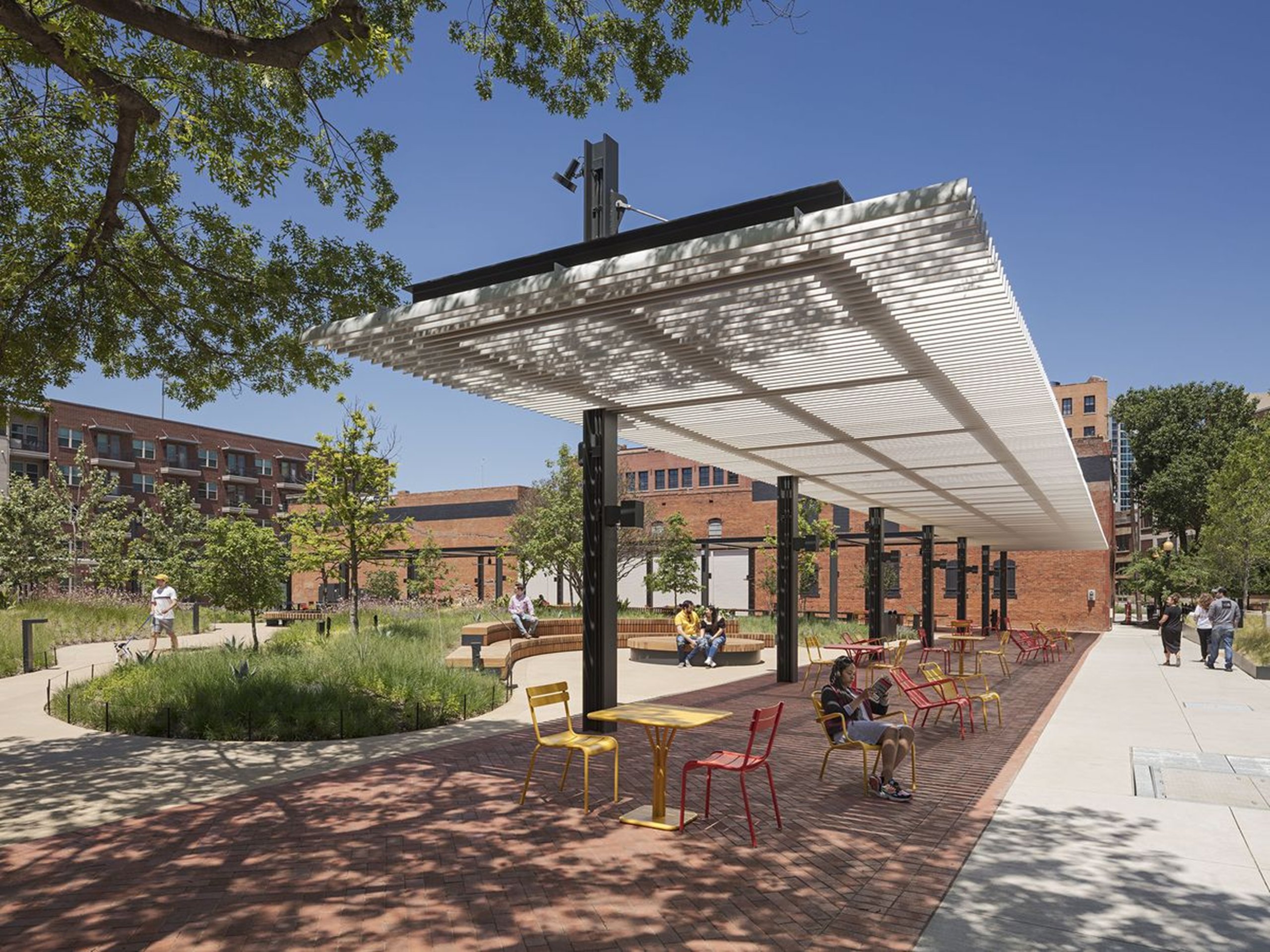 Outdoor seating area with colorful chairs and tables under a white slatted canopy in an urban park.