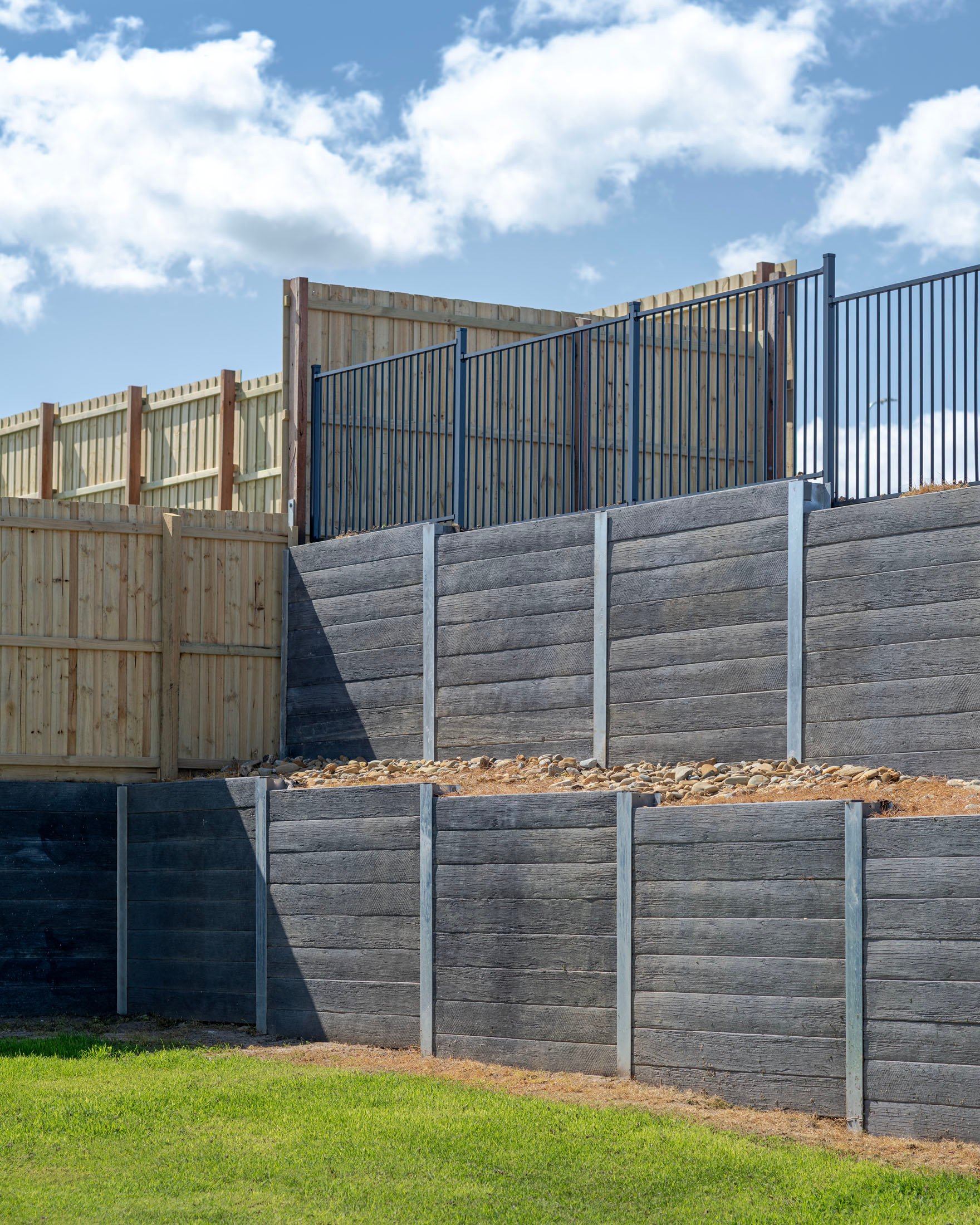 Terraced wooden retaining walls with metal supports and fences under a blue sky with clouds.