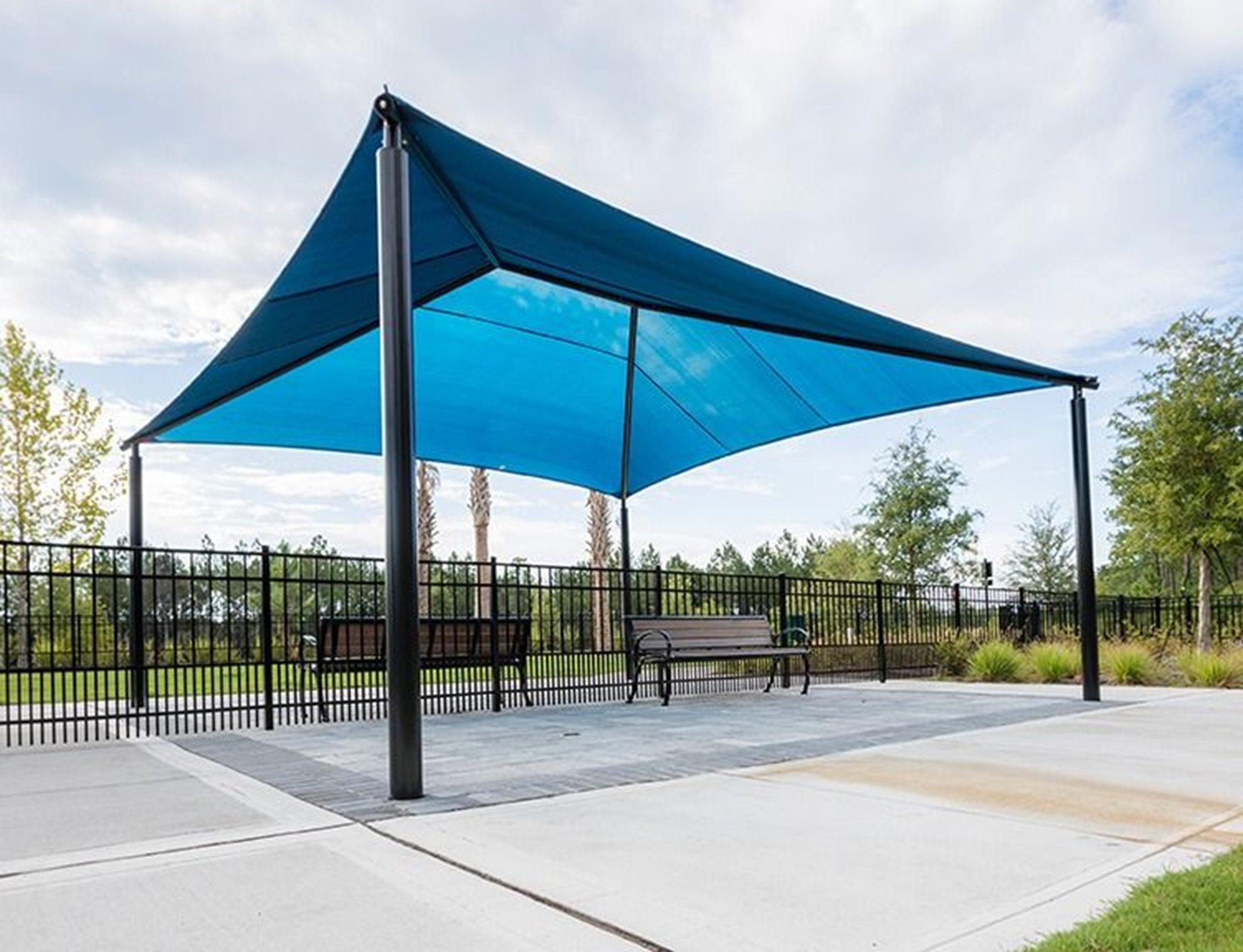 Blue triangular shade structure with black poles over benches on paved ground in a park area.