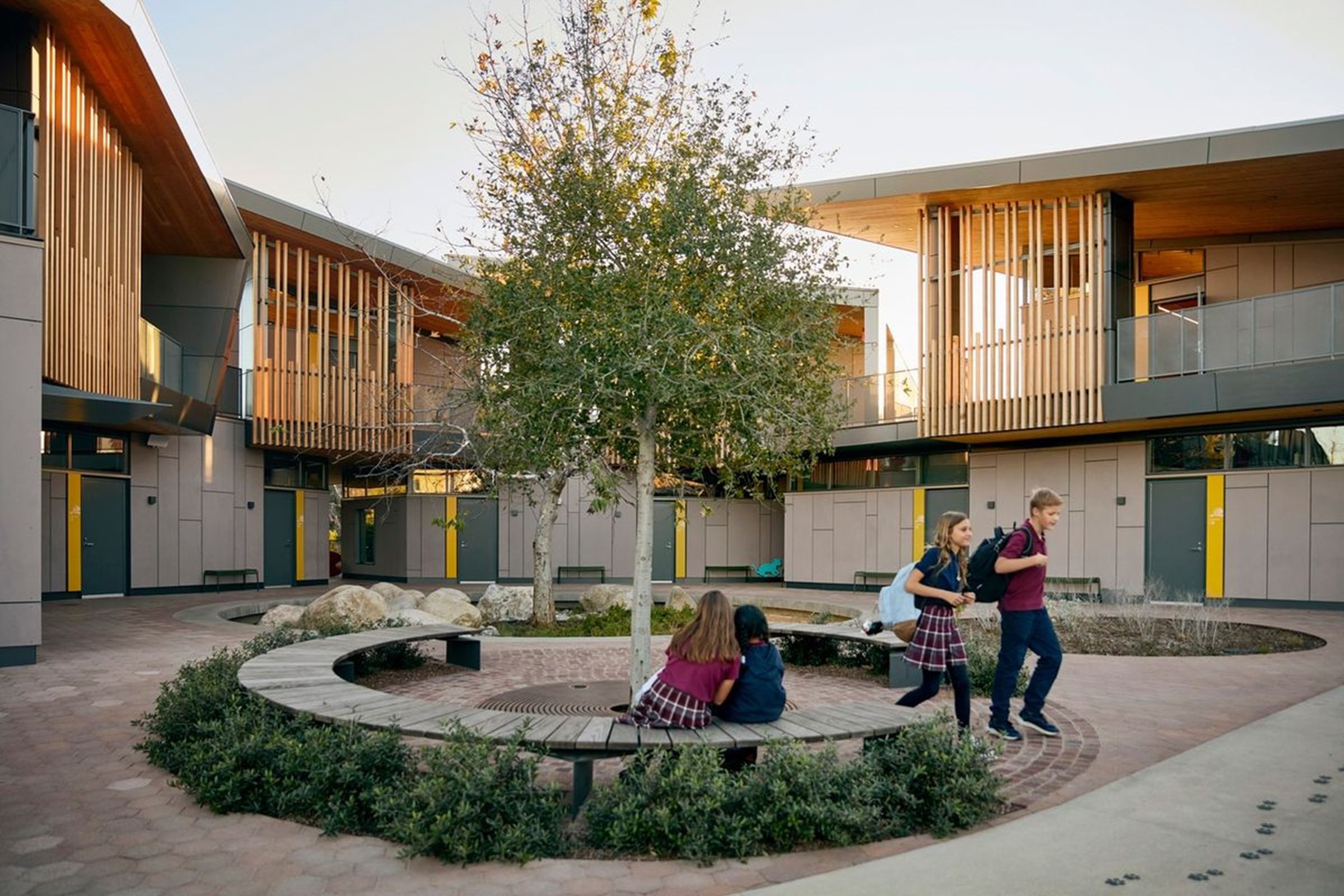 Modern school courtyard with circular bench around tree and children walking and sitting nearby.