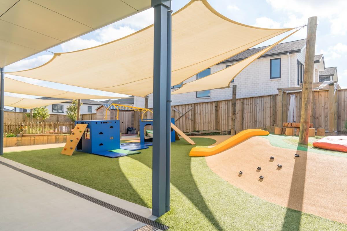 Outdoor playground with climbing structures, a slide, and shade sails in a fenced yard.