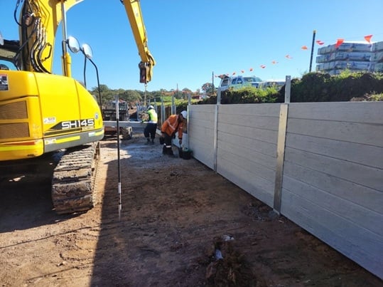 Workers installing concrete panels along a dirt pathway with a yellow excavator on site.