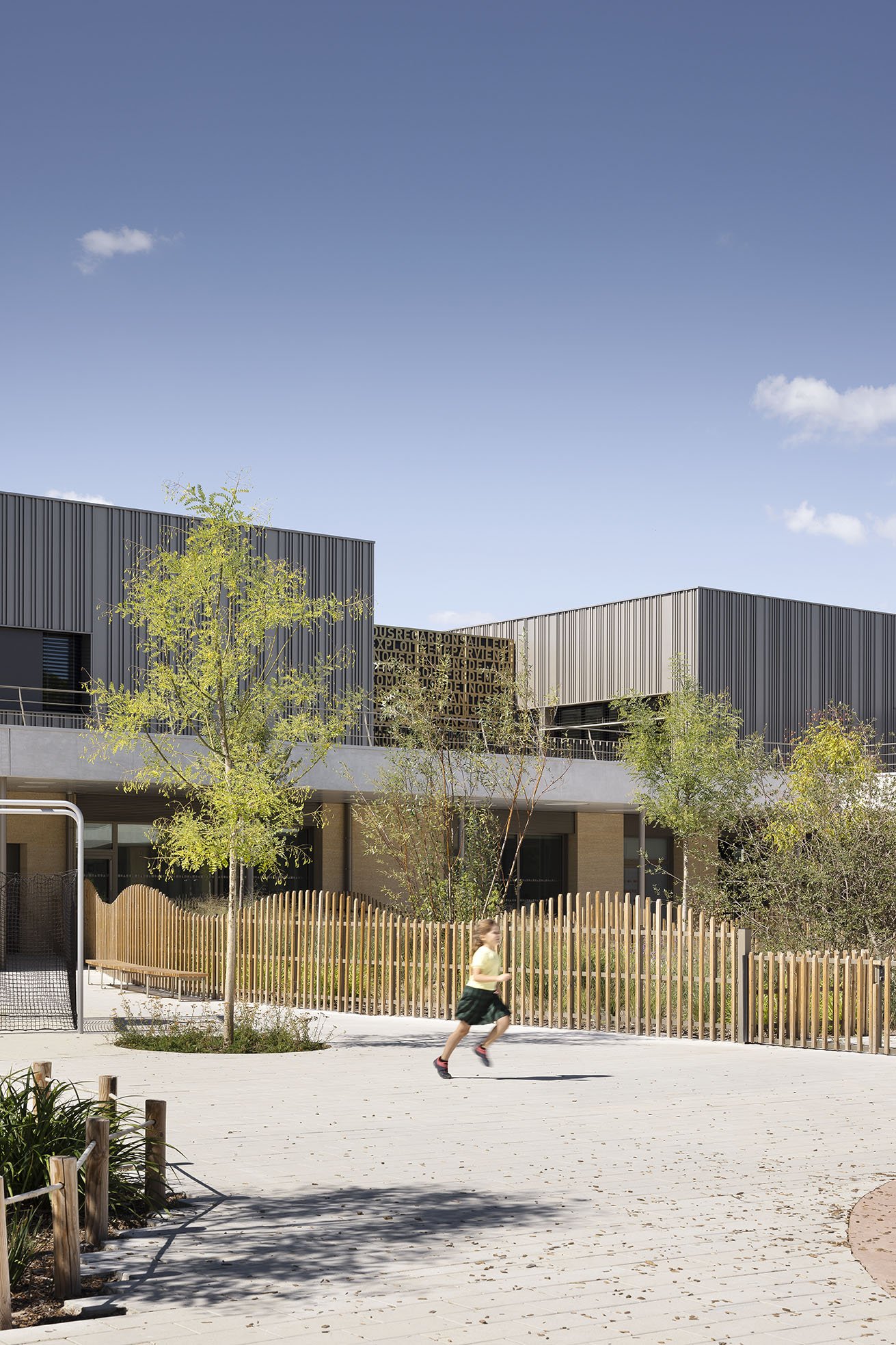 Child running on paved courtyard with trees, wooden fence, and modern building in background under clear sky.