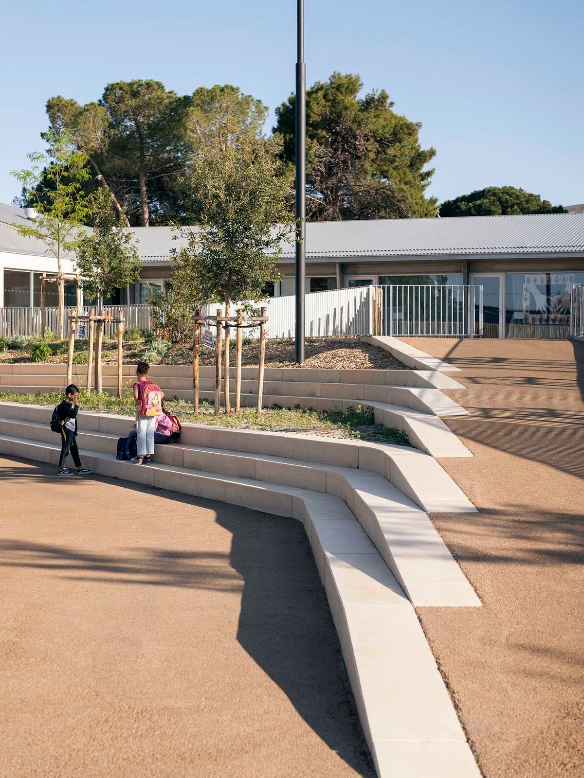Two children with backpacks near terraced concrete seating and small trees in an outdoor school area.
